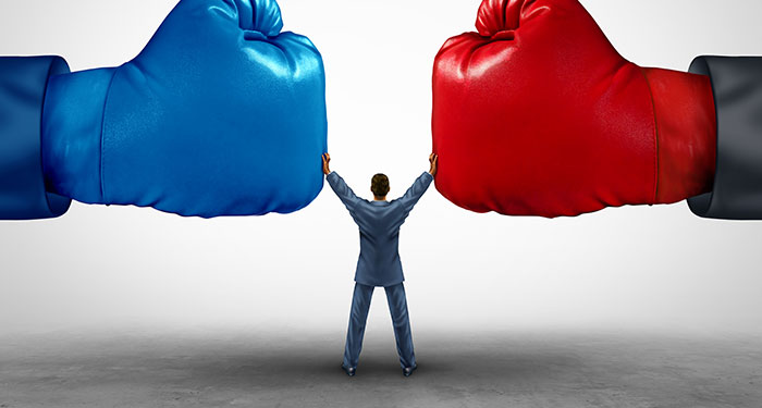A person in a suit standing between two giant red and blue boxing gloves holding his hands up to stop them